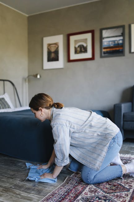 A woman engaged in surface cleaning using a yellow vacuum cleaner with a long hose attachment on a patterned area rug. She is wearing casual clothing, including a beige shirt and blue jeans, and is fo