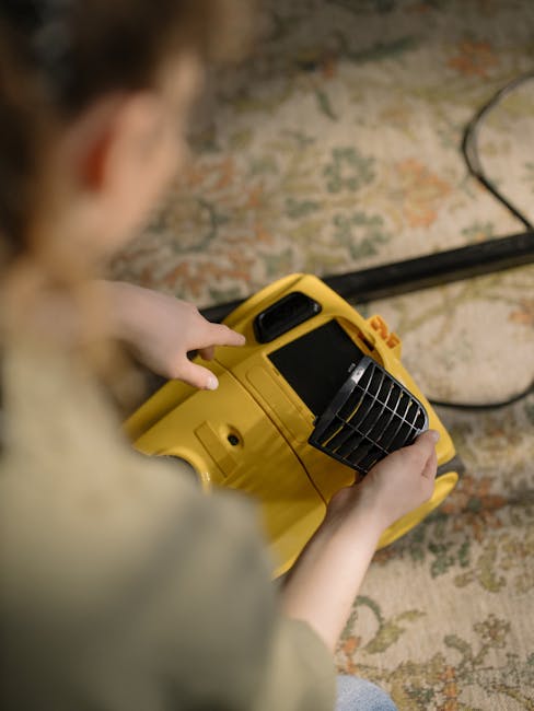 A person is holding a yellow portable air mover or fan with adjustable vents, placed on a patterned, multi-colored carpet in a room. The individual, whose face is not visible, is operating the device to facilitate surface cleaning or drying. The room features a textured wall with a floral or abstract pattern and is illuminated by natural or ambient lighting. The scene emphasizes the importance of thorough cleaning, ventilation, or drying processes in domestic or commercial environments, as part of professional surface or deep cleaning services offered by Carpet Cleaning Greenwich.