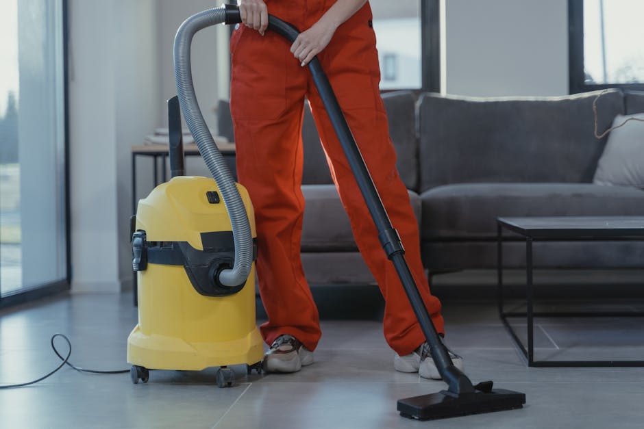 A person is holding a yellow portable air mover or fan with adjustable vents, placed on a patterned, multi-colored carpet in a room. The individual, whose face is not visible, is operating the device to facilitate surface cleaning or drying. The room features a textured wall with a floral or abstract pattern and is illuminated by natural or ambient lighting. The scene emphasizes the importance of thorough cleaning, ventilation, or drying processes in domestic or commercial environments, as part of professional surface or deep cleaning services offered by Carpet Cleaning Greenwich.