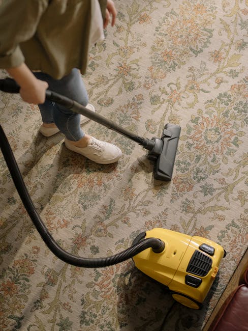 A person is performing domestic cleaning on a patterned area rug in a living room using a yellow vacuum cleaner with a black hose attachment. The individual, wearing light-colored sneakers, jeans, and a beige coat, is holding the vacuum's wand and moving it across the surface. The rug features floral motifs in shades of beige, green, and orange, and is positioned on a wooden floor. Bright lighting illuminates the space, highlighting the clean and well-maintained appearance of the rug. This deep cleaning process exemplifies surface cleaning and sanitisation services offered by Carpet Cleaning Greenwich, aimed at maintaining hygiene and aesthetic appeal of residential interiors.