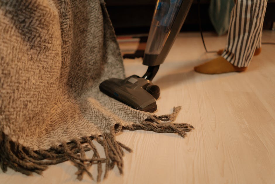 A yellow canister vacuum cleaner with a long black hose and cleaning head is positioned on an ornate patterned area rug in a living room with wooden flooring. To the left, a black low-profile TV stand