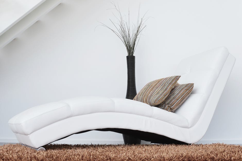 A modern white chaise lounge with a smooth, leather surface is positioned in a bright, minimalistic living room with white walls and a soft brown carpet. The chaise is decorated with two striped cushions in neutral colors. Behind the lounge, there is a tall black vase containing slender, dried decorative grasses, adding an elegant touch to the space. The room features clean lines and natural lighting, emphasizing a tidy and hygienic environment suitable for surface cleaning, deep cleaning, and sanitisation as part of domestic cleaning services offered by Carpet Cleaning Greenwich near Cutty Sark Greenwich. The setup reflects a well-maintained interior prepared for effective cleaning and hygiene maintenance.
