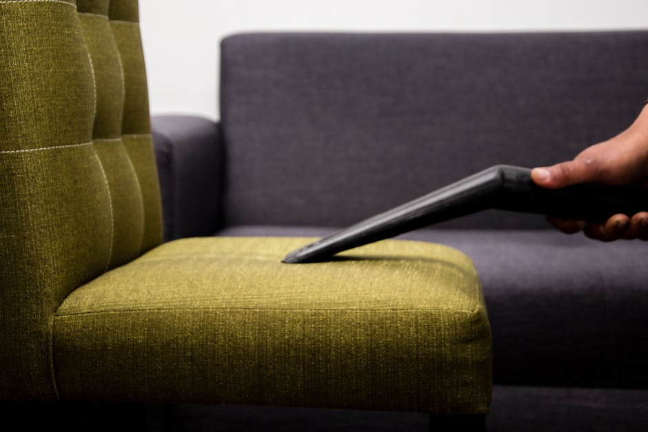 Close-up of a person using a handheld vacuum cleaner to deep clean a green upholstered armchair in a living room setting, with a grey fabric sofa visible in the background. The upholstery appears well-maintained and free of visible dust or debris, with the scene illuminated by natural or soft indoor lighting. The cleaning process emphasizes surface cleaning and hygiene, consistent with professional upholstery cleaning services offered by Carpet Cleaning Greenwich near Cutty Sark Greenwich, as highlighted on their website.