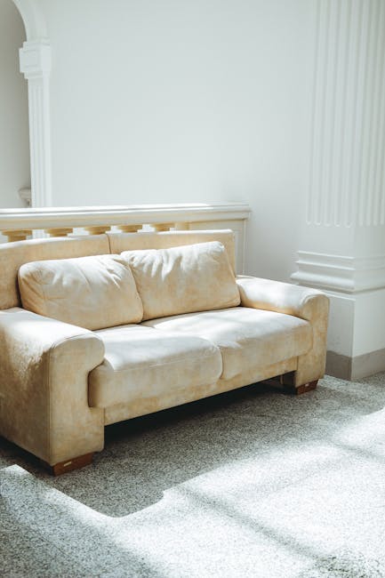 A modern white chaise lounge with a smooth, leather surface is positioned in a bright, minimalistic living room with white walls and a soft brown carpet. The chaise is decorated with two striped cushions in neutral colors. Behind the lounge, there is a tall black vase containing slender, dried decorative grasses, adding an elegant touch to the space. The room features clean lines and natural lighting, emphasizing a tidy and hygienic environment suitable for surface cleaning, deep cleaning, and sanitisation as part of domestic cleaning services offered by Carpet Cleaning Greenwich near Cutty Sark Greenwich. The setup reflects a well-maintained interior prepared for effective cleaning and hygiene maintenance.